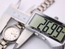 A digital caliper precisely measuring the watch case diameter at 26.99mm, with the watch visible in the background.