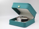 The Mondaine watch displayed inside a generic green leatherette studio presentation box with a gold crown emblem.