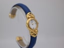 Another angled view of the watch, emphasising the intricate gold-tone bow-like lugs and the contrasting blue leather of the strap, presented on a clear stand.
