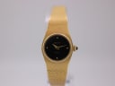 Front view of the gold-tone Zenith quartz watch on a clear display stand, showing the black dial and integrated mesh bracelet.