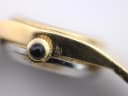 Close-up of the gold-tone watch case side showing the black cabochon crown and an embossed maker's mark.