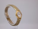 Another angled side view of the gold-tone Seiko watch on a clear stand, highlighting the bracelet texture and case profile.