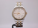 Front view of the Tissot Ballade two-tone watch, showcasing the cream dial with gold Roman numerals and hands on a watch stand.