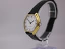 Side profile view of the Bulova Longchamp watch on a stand, highlighting the gold case and crown, with the black leather strap curving behind.