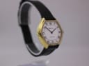 Angled side view of the Bulova Longchamp watch on a clear stand, showing the gold case, white dial, and black leather strap from the other side.