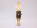 Front view of the gold-tone Orient quartz watch on a clear stand, showing the black rectangular dial and integrated bracelet.
