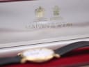 Close-up of open Mappin & Webb original presentation box with gold crests and logo, watch visible out of focus in foreground.