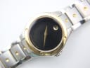 Close-up of the Movado SE watch dial, showing the black face, gold museum dot, and gold hands on a white background.