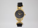 Front view of the vintage Gucci 6000 L watch on a clear display stand, showing the black dial and gold-tone case.