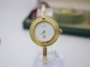 Front view of the Gucci 1100-L watch on a clear display stand, showing the white dial with gold baton hands and an elegant gold-tone bezel.