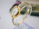 Angled side view of the Gucci 1100-L watch on a clear display stand, highlighting the integrated gold-tone bangle bracelet.