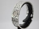 Angled view of the Gucci 112 watch showing the polished silver case and engraved 'G' monogram on the bangle, on a black stand.