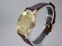 Angled side view of the gold tone Gucci 5100 M watch, showing the crown and case contours, on a clear display stand.
