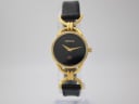 Front view of the Gucci 6000L watch on a clear display stand, showing the black dial, gold-tone case, and black leather strap.
