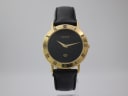 Front view of the Gucci 3000 M watch on a clear stand, showing its black dial, gold-tone case, and black lizard-effect leather strap.