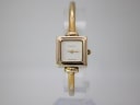 Front view of the Gucci 1900 L watch on a clear stand, showing its square gold-tone case and white dial with minimalist hands.