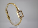 Side profile view of the Gucci 1900 L watch on a clear stand, detailing the elegant curve of the gold-tone bangle.