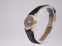 Angled side view of the Raymond Weil watch on a clear stand, highlighting the gold electroplated case and crown