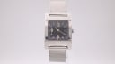 Front view of the GUCCI 7700L watch with a silver rectangular case and black dial, on a reflective white surface.