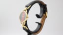 Side-angle view of the Gucci watch on a display stand, highlighting the gold-tone case and black leather strap.