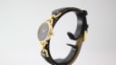 Side view of the Gucci 6000L watch on a stand, showcasing the profile of the gold-toned case and the black leather strap.