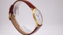 Another side profile view, showing the gold-plated case and crown, with the brown leather strap on a clear display stand.