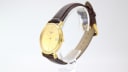 Angled side view of the Tissot watch showing the gold-tone case and the brown leather strap on a display stand.