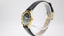 Side-angled view of the Amara watch on a clear display stand, highlighting the gold-tone case and textured black leather strap.