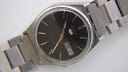 Angled close-up view of the black dial with gold baton indices and hands, showing the day and date window.
