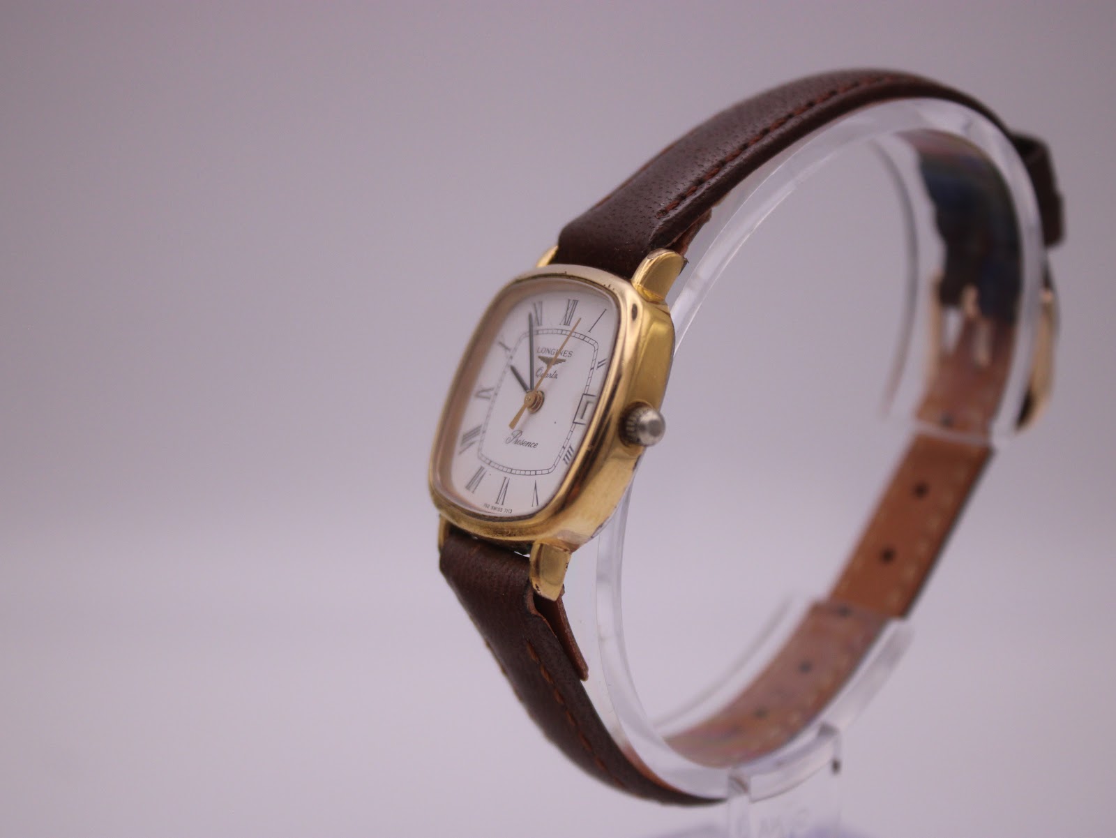 Angled view of Longines Presence watch showing side of gold case and brown leather strap on clear display stand.