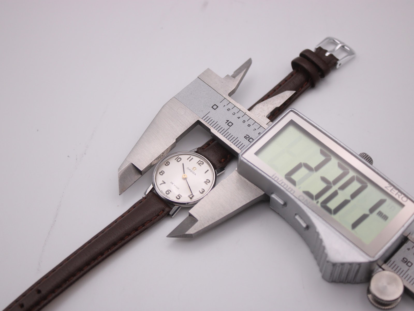 Overhead view of the Omega De Ville watch being measured by a digital caliper, displaying a case size of 23.07mm.