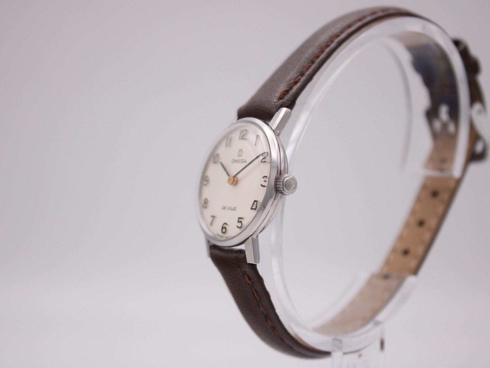 Angled side view of the Omega De Ville watch, highlighting its slender profile and brown leather strap on a watch stand.