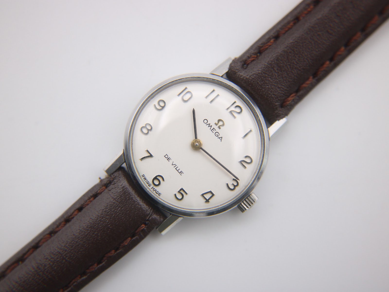 Close-up front view of the Omega De Ville watch dial, showing the cream face, black Arabic numerals, and gold-tone hands.