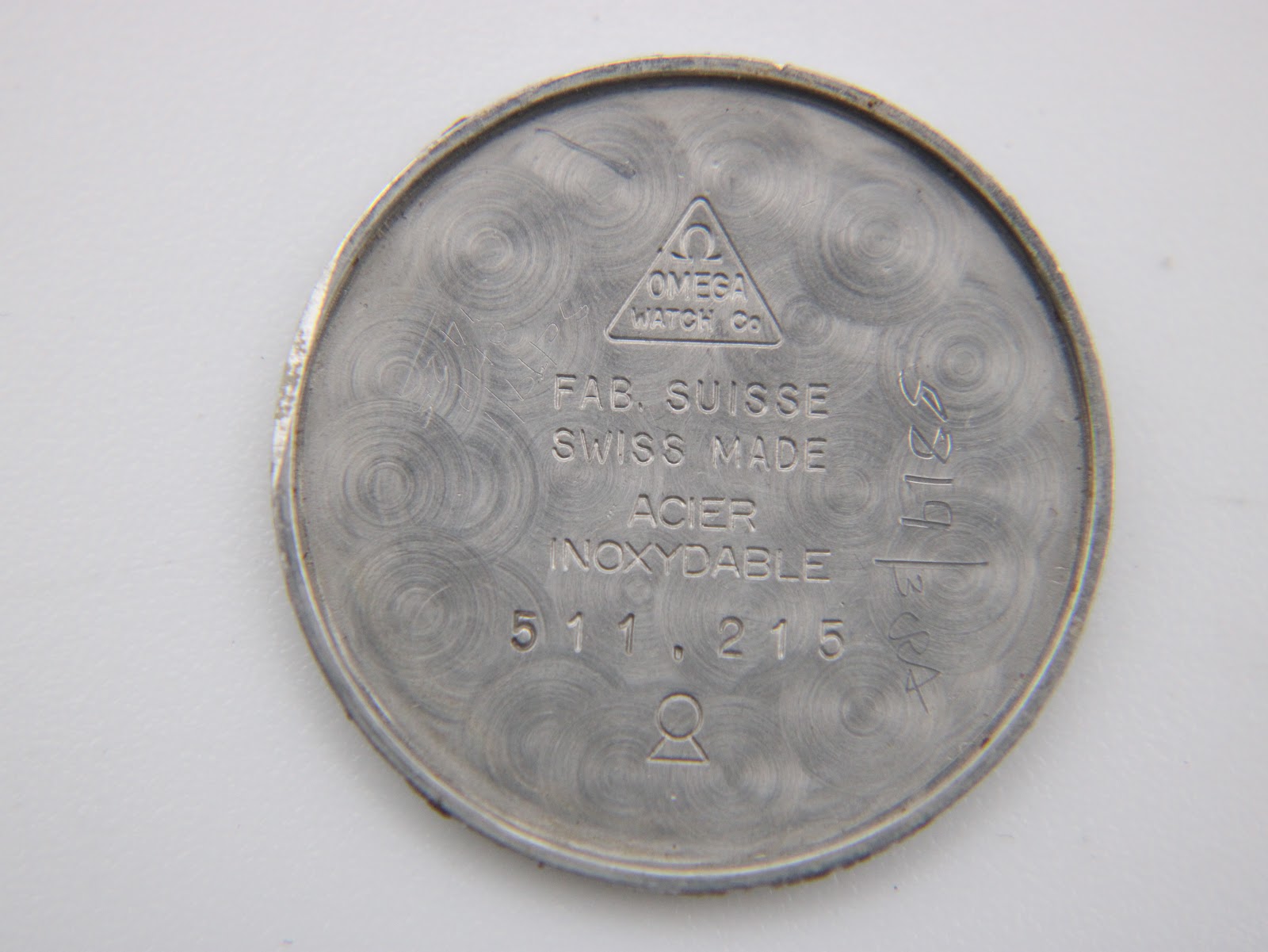 Close-up of the removed stainless steel case back, showing 'OMEGA WATCH Co', 'FAB. SUISSE', 'SWISS MADE', 'ACIER INOXYDABLE', and model number '511,215'.
