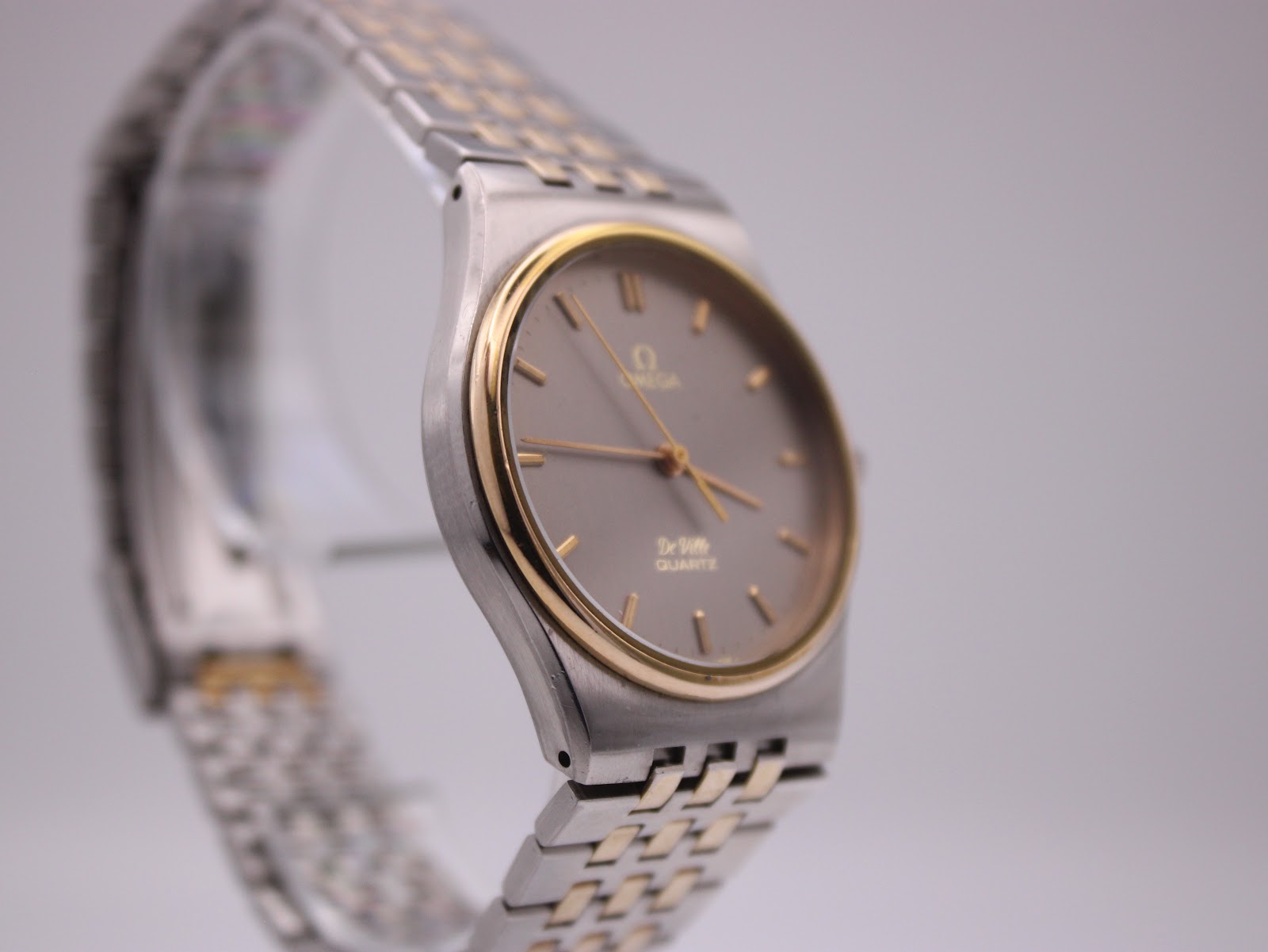 Angled view of the Omega De Ville watch, highlighting the gold-plated bezel and grey dial.