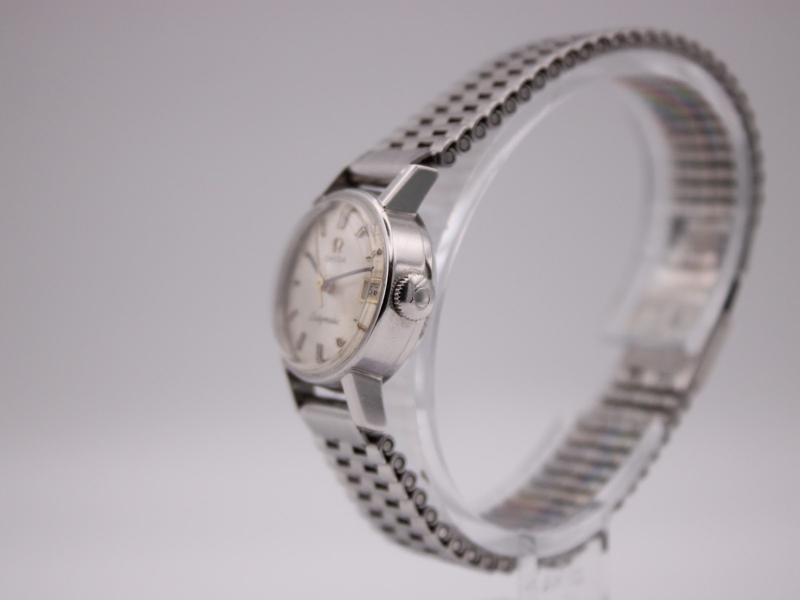 Side profile view of the Omega Ladymatic watch on a clear stand, showing the signed crown and steel bracelet.