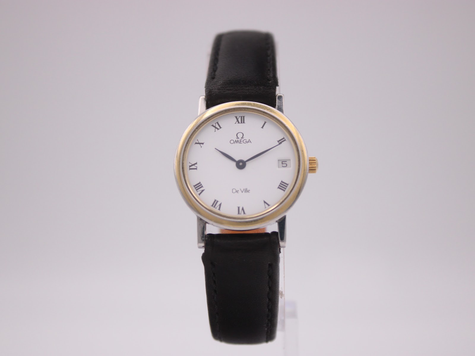 Front view of the Omega De Ville two-tone quartz watch with white Roman numeral dial, on a clear display stand.
