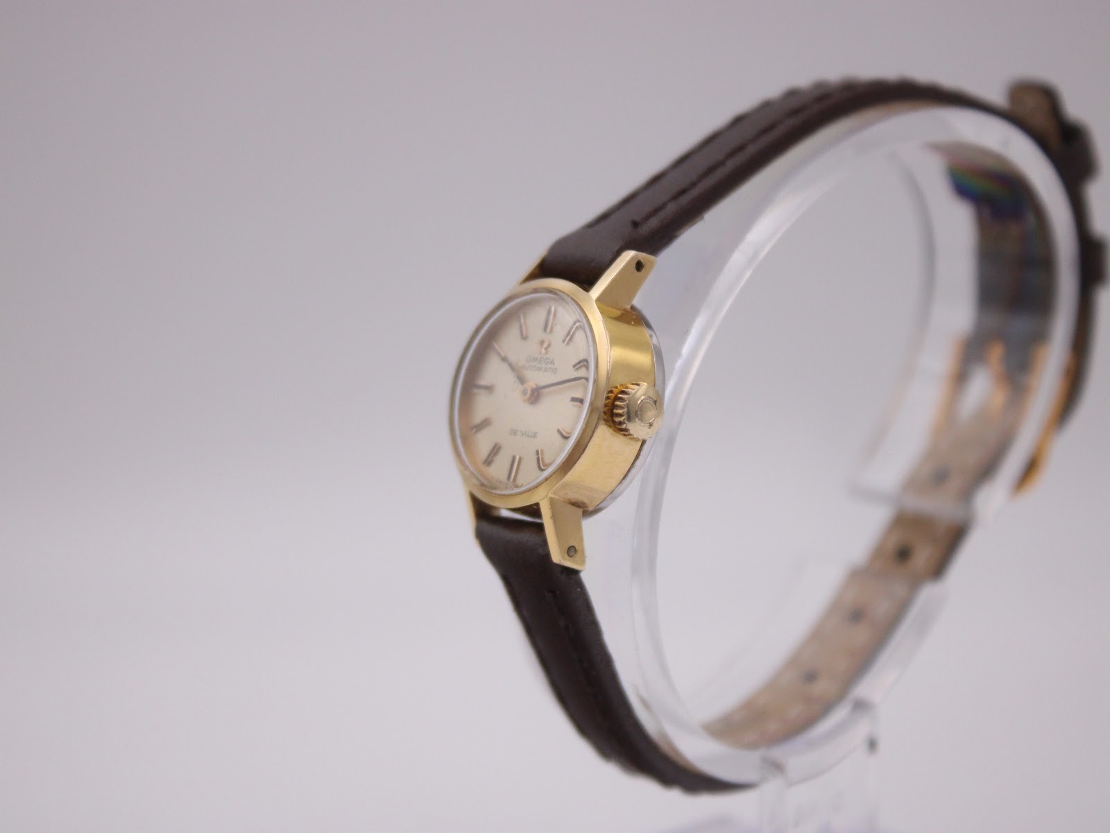 Side profile view of the Omega De Ville watch on a stand, highlighting the gold-plated case and crown.