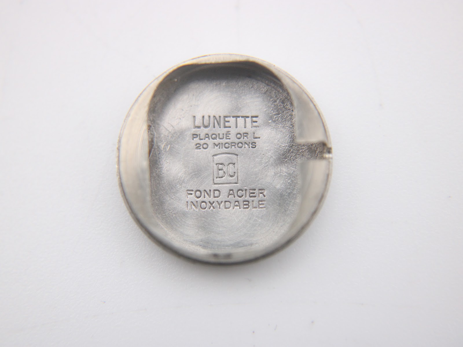 Internal view of the watch case back, engraved with 'LUNETTE PLAQUÉ OR L. 20 MICRONS' and 'FOND ACIER INOXYDABLE'.