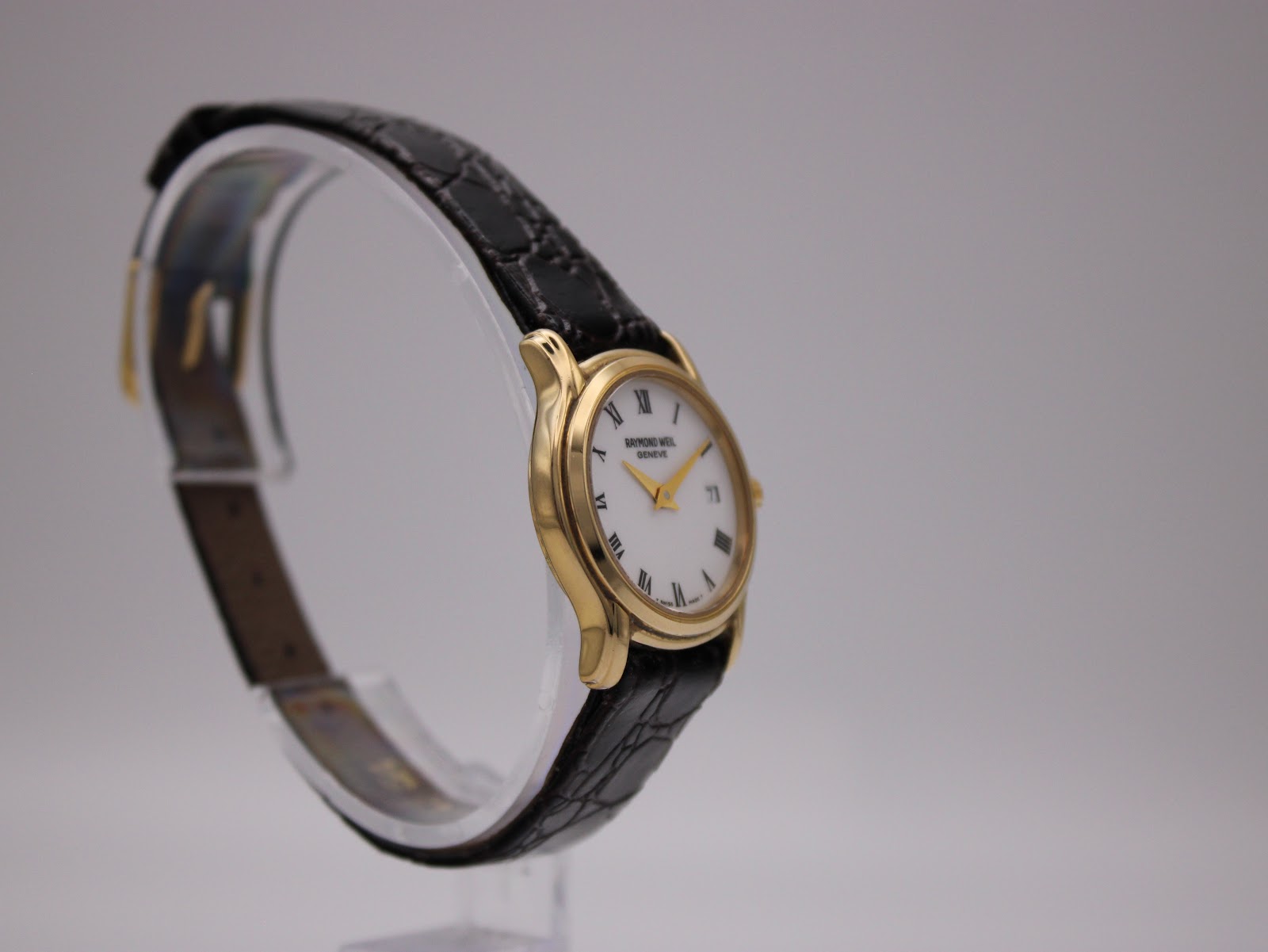 Angled view of the Raymond Weil watch highlighting the gold case and textured black leather strap on a clear stand.