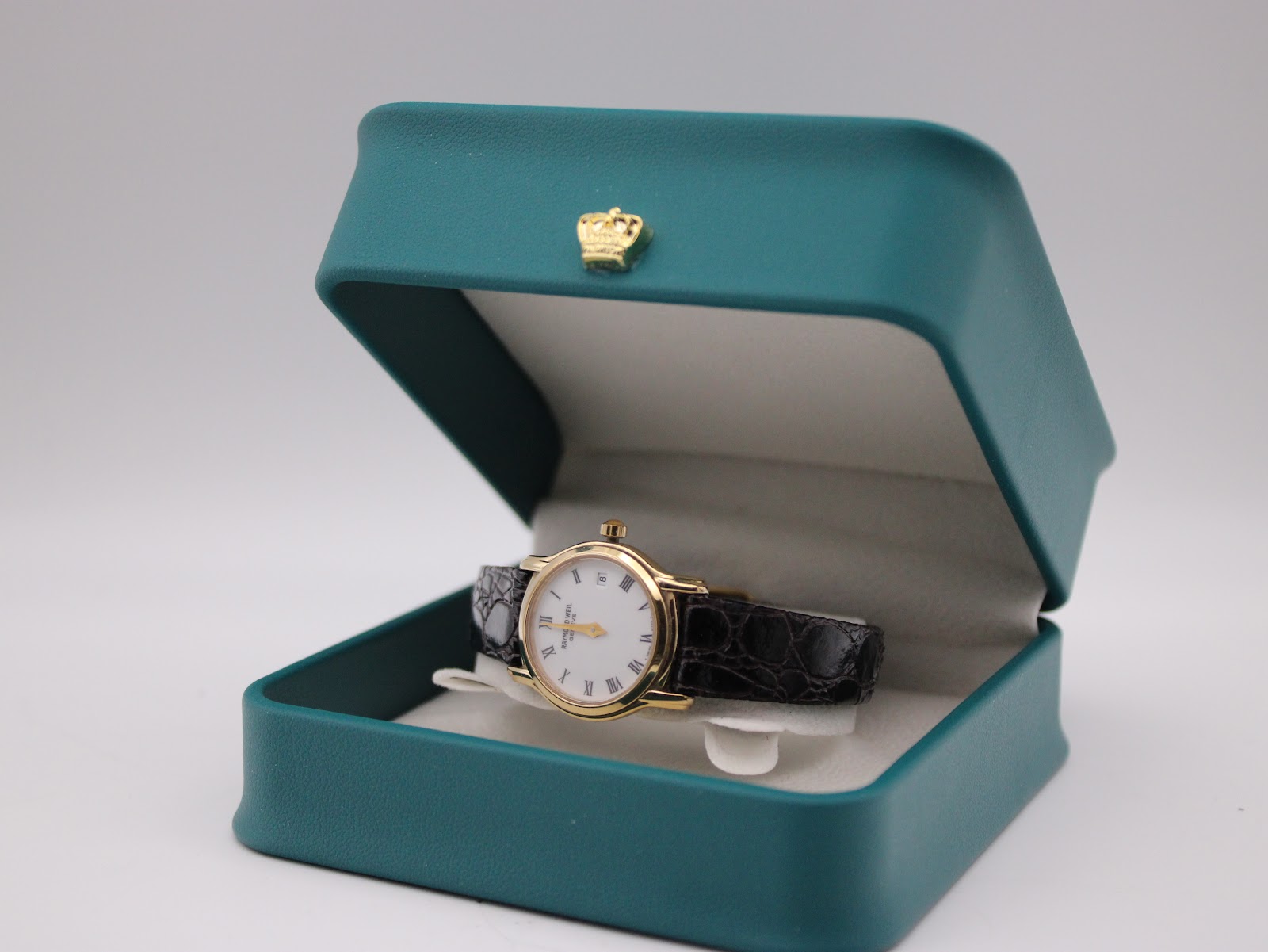The Raymond Weil watch resting inside its original teal presentation box with a gold crown logo on the lid.