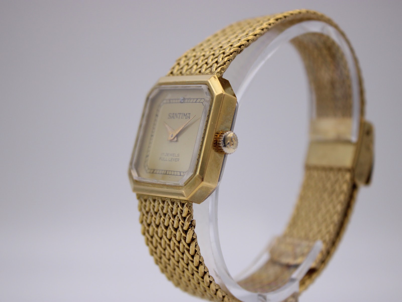 Angled view of Santima watch on a clear stand, showing the signed crown and side profile.
