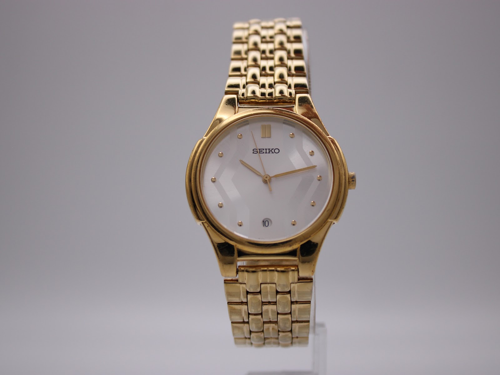 Front view of the Seiko V732-0450 gold-tone quartz watch with white dial, showing textured pattern and date window.