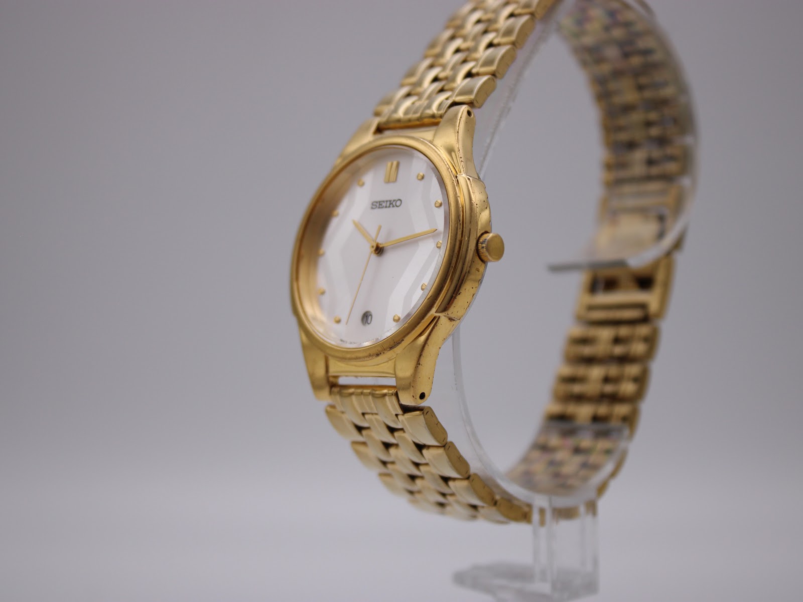 Angled side view of the Seiko V732-0450 watch on a clear stand, highlighting the gold-tone case and integrated bracelet.