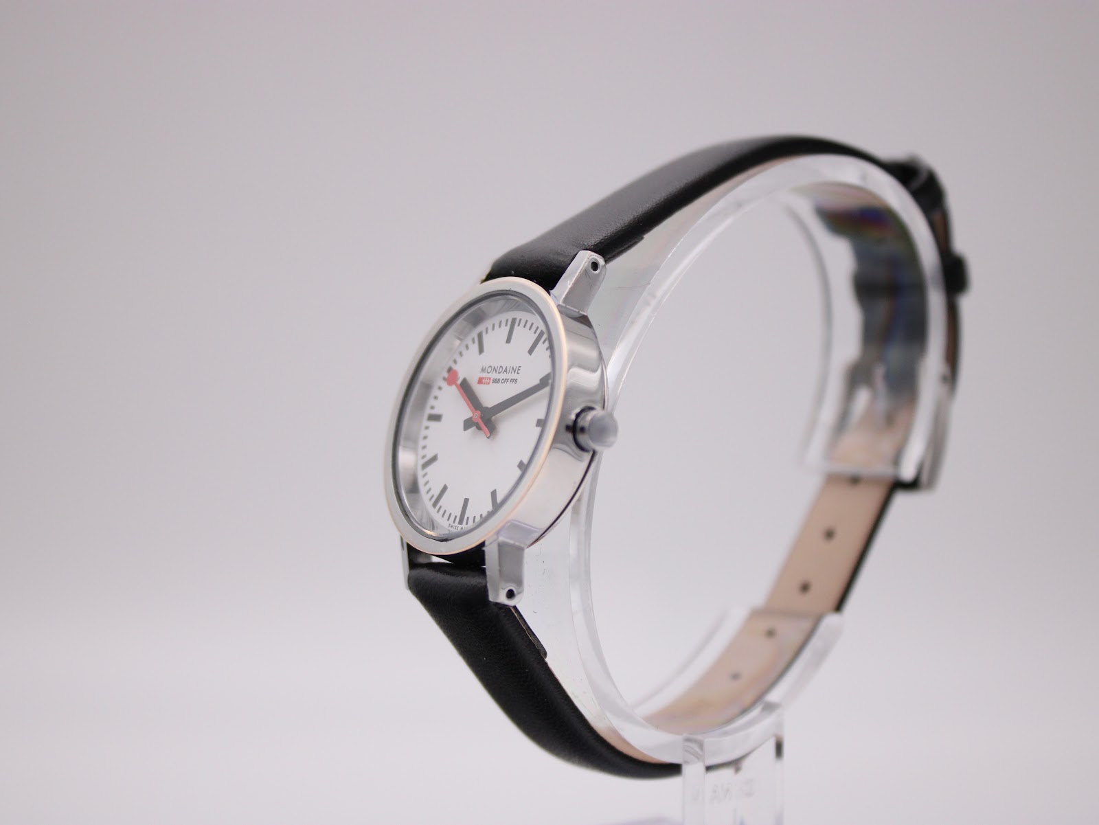 Angled side view of the Mondaine watch showing the silver case profile and black leather strap on a clear stand.