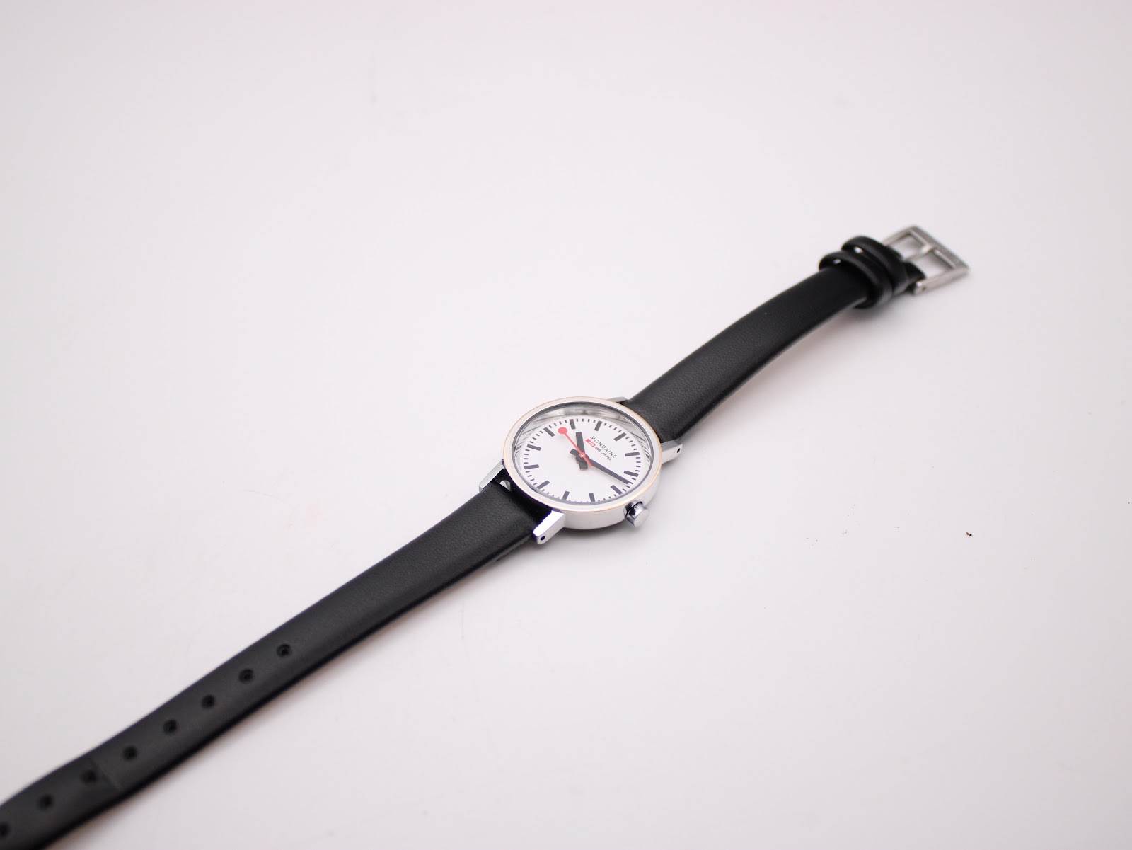 Overhead angled view of the Mondaine watch, showing the full length of the black leather strap against a white background.