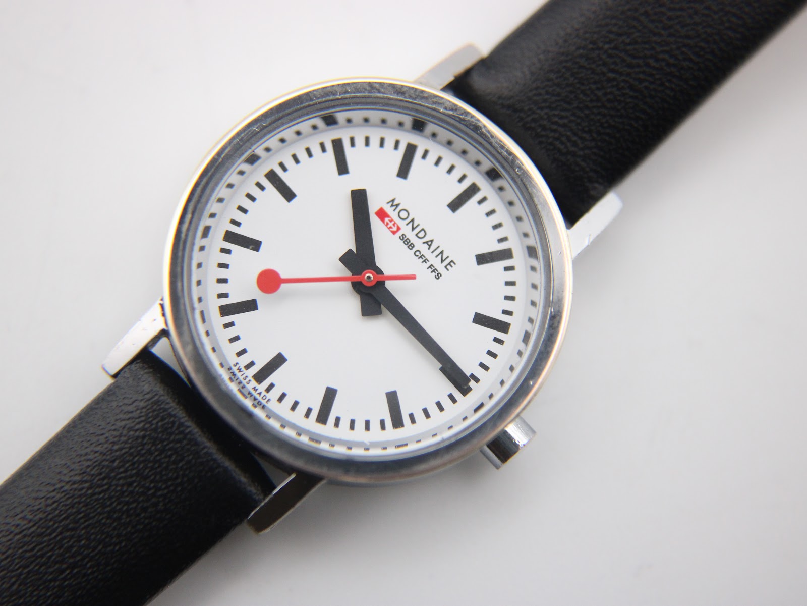 Close-up of the Mondaine watch dial, highlighting the iconic black baton markers and red lollipop seconds hand.