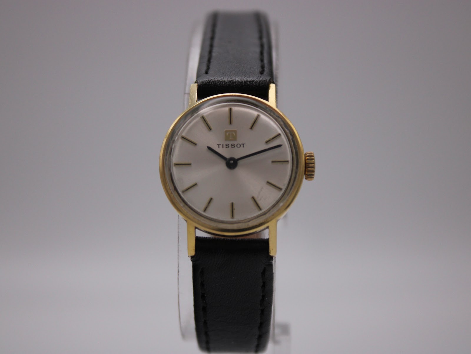 Front view of vintage Tissot ladies' watch on a display stand, showing gold case and silver dial with black leather strap.