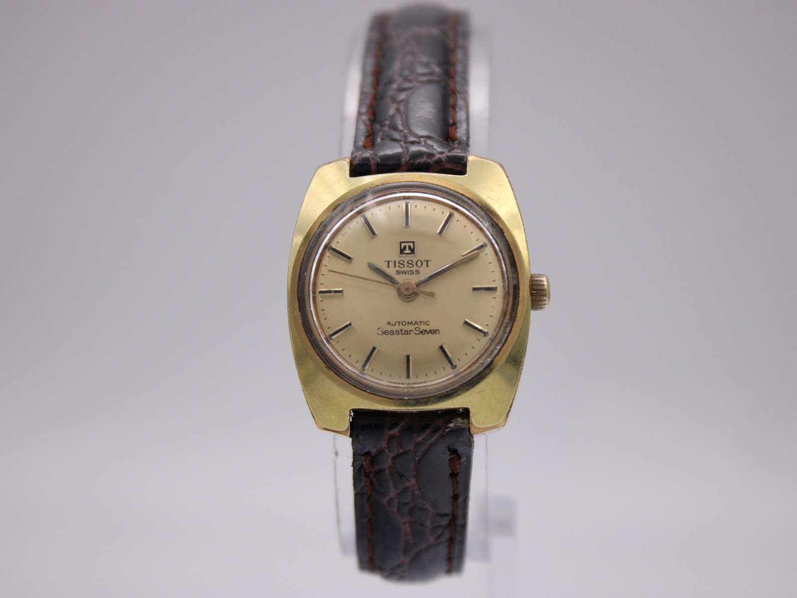 Front view of gold-tone Tissot Seastar Seven with champagne dial and brown leather strap on a watch stand.