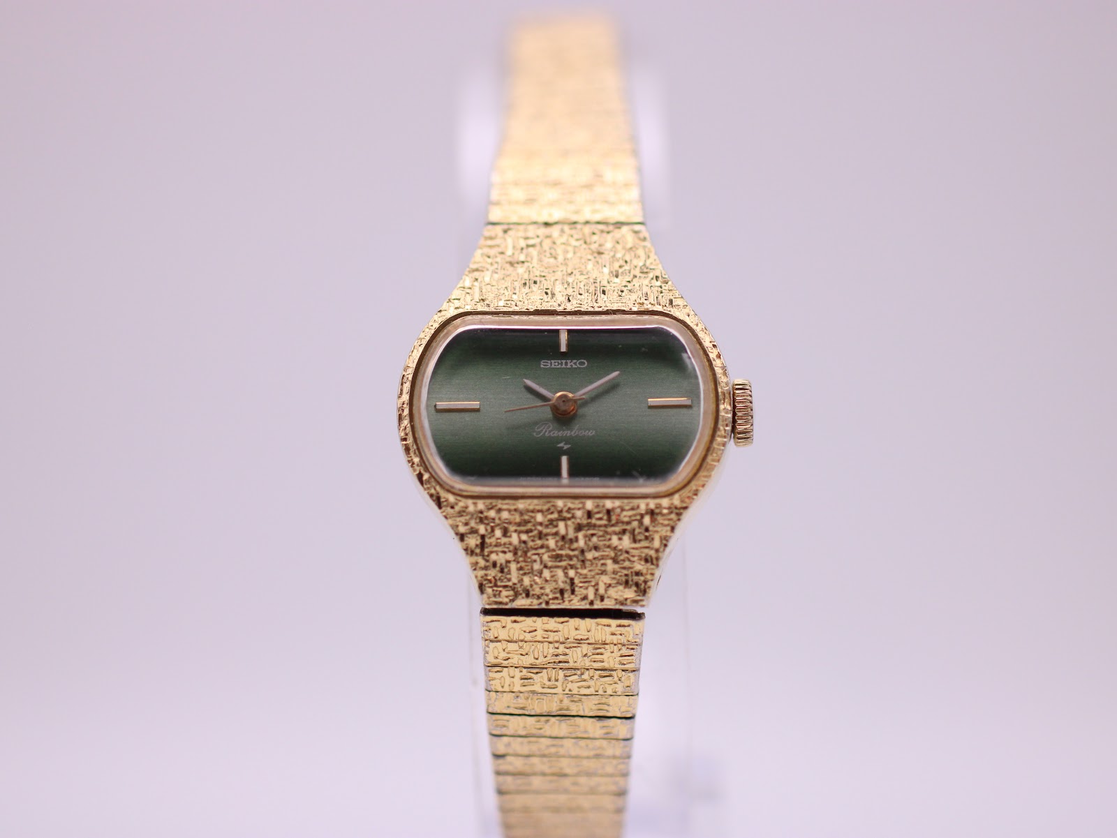 Front view of the Seiko Rainbow watch with a green dial and gold-tone rectangular textured case on a display stand.